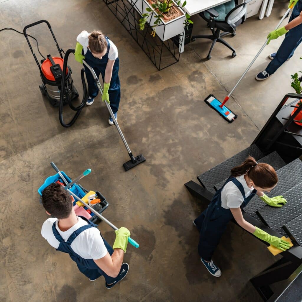 Équipe de quatre agents d'entretien professionnels nettoyant un bureau moderne. Ils utilisent aspirateur, serpillière et chiffons pour sol et escalier.