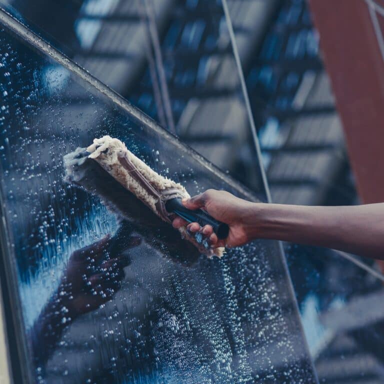 Main nettoie une grande vitre avec un laveur. Le verre est couvert de mousse savonneuse, avec des reflets bleutés.